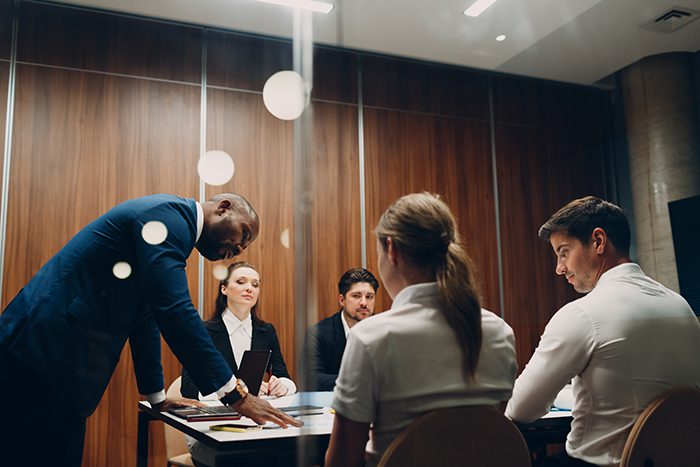 A man in a suit presents documents to four colleagues seated around a conference table in a modern office meeting room.