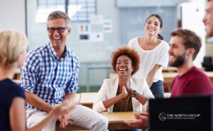 A group of six adults sits and stands around a table, smiling and laughing in a bright, modern office space.