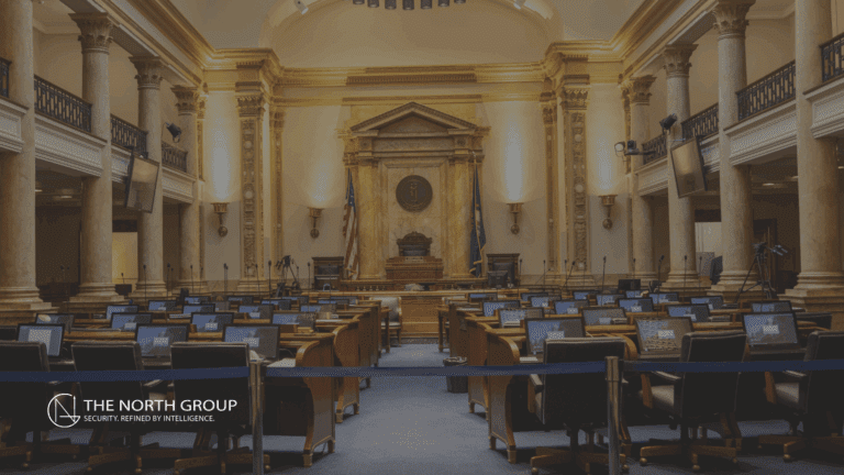 A large, empty legislative chamber with rows of desks, computer monitors, and columns—a solemn space addressing issues like violence against elected officials—features an American flag and a judge’s bench at the front.