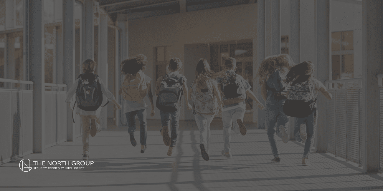 A group of school children with backpacks run across a pedestrian bridge toward a Safe School building on a sunny day. The North Group logo appears in the bottom left corner.