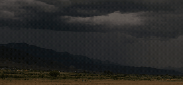 Dark storm clouds gather over distant mountains, casting shadows on a flat, rural landscape with sparse vegetation and small buildings.