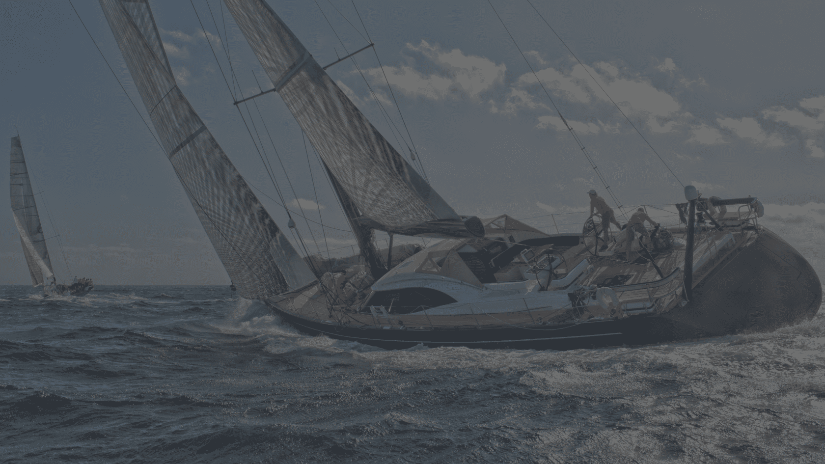 A sailboat with several crew members navigates choppy ocean water under partly cloudy skies, with another sailboat visible in the distance.