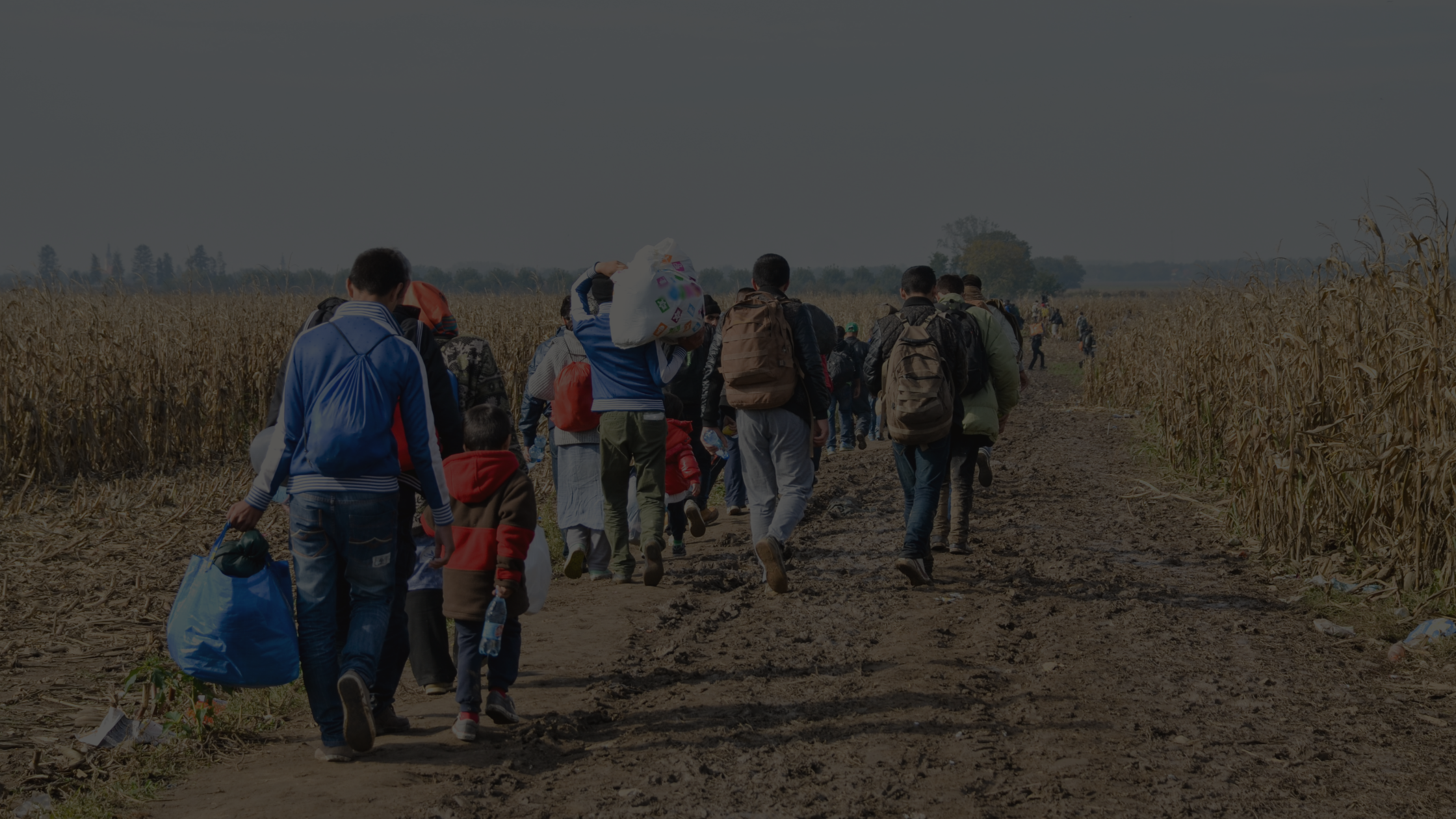 A group of people, including children, walk along a muddy rural path carrying bags and backpacks through a field.