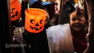 Children in Halloween costumes hold out orange pumpkin and white bags while trick-or-treating, smiling and looking excited. The North Group logo appears in the corner.