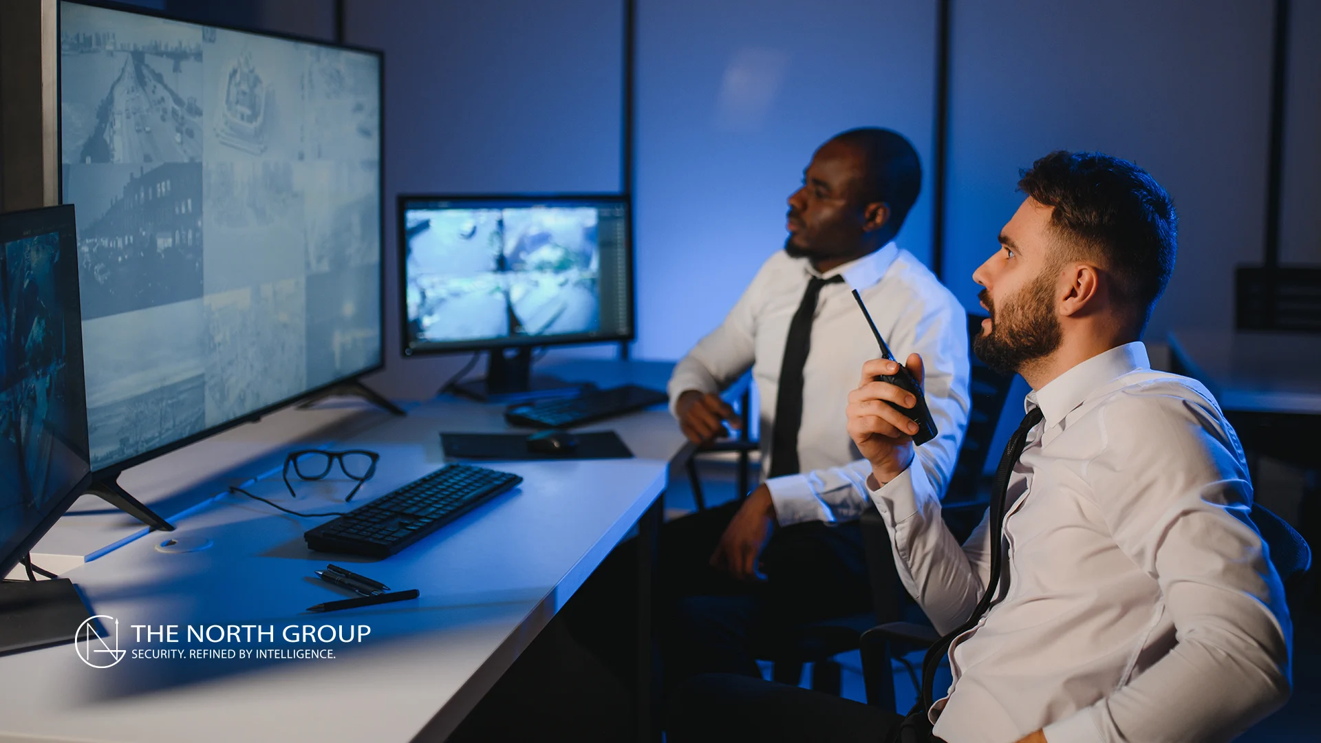Two men in business attire monitor multiple security camera feeds on computer screens in a control room. One man holds a walkie-talkie. The North Group logo is visible on the desk.