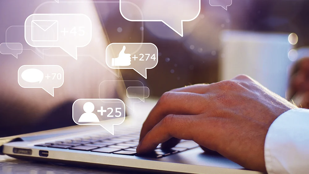 A person types on a laptop keyboard with digital icons above showing message, like, and comment notifications.