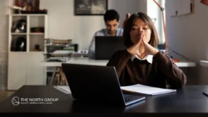 A woman sits at a desk with a laptop and notebook, hands pressed together thoughtfully, while a man works in the background.