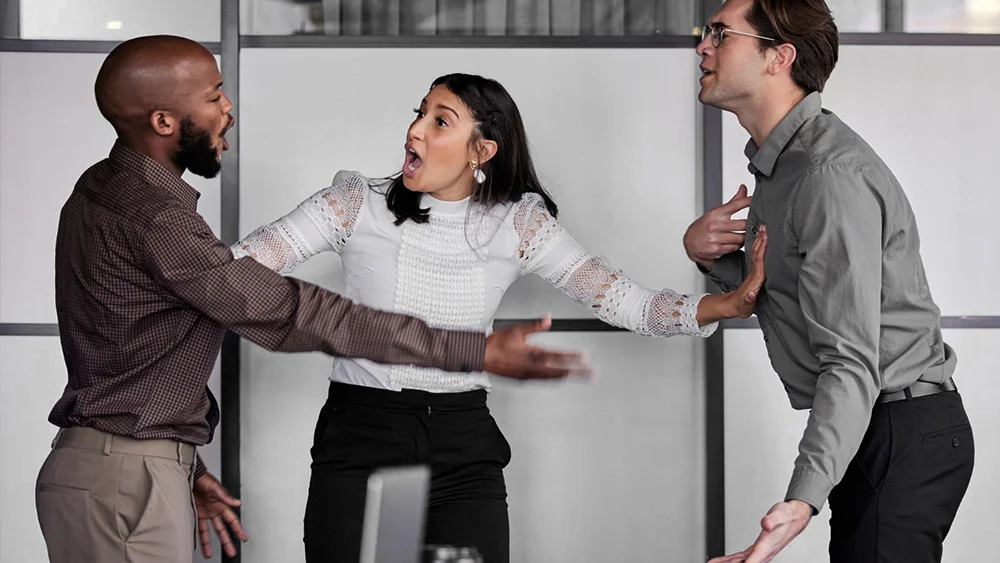 Three people in business attire are having a heated argument in an office setting; a woman stands between two men, gesturing to separate them.