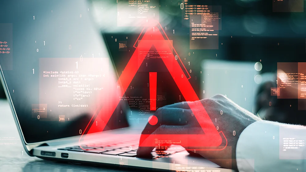 A person uses a laptop with a large red warning symbol overlay, highlighting a cybersecurity threat that calls for immediate protective intelligence measures.
