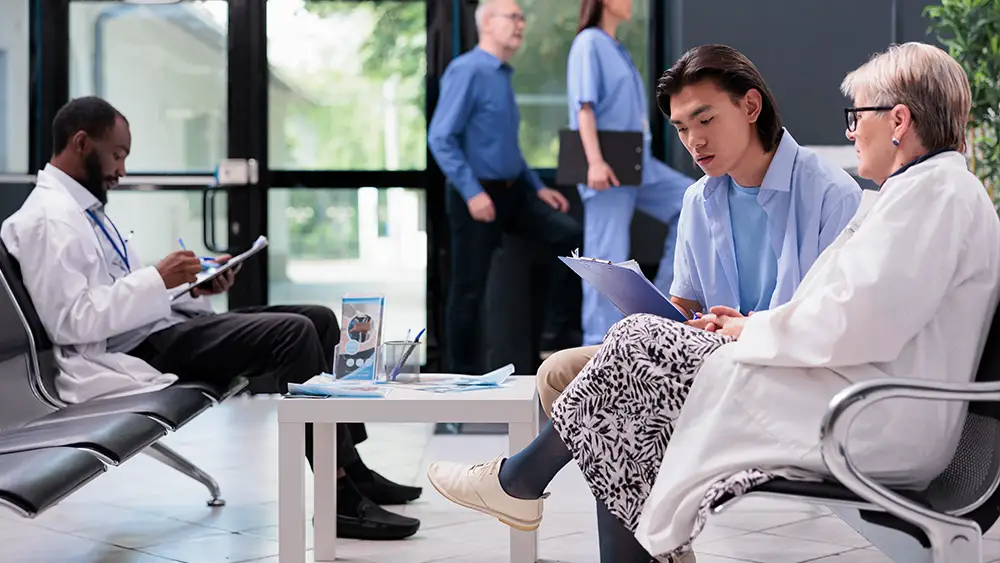 Medical professionals sit and review documents in a waiting area, while two others walk by in the background. The setting appears to be a modern healthcare facility.