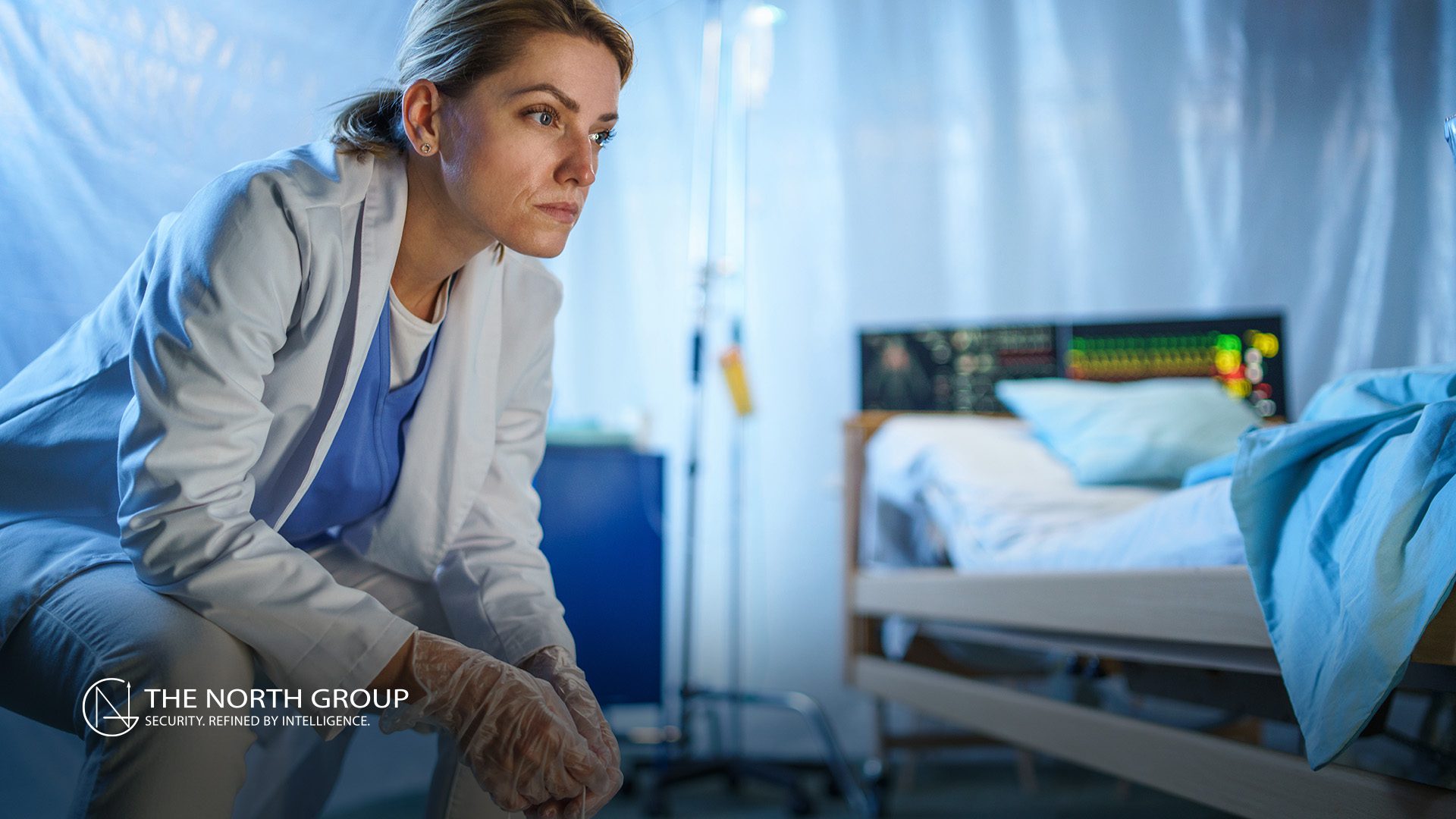 A healthcare professional in scrubs and gloves crouches beside a hospital bed in a medical setting, with monitoring equipment visible in the background.