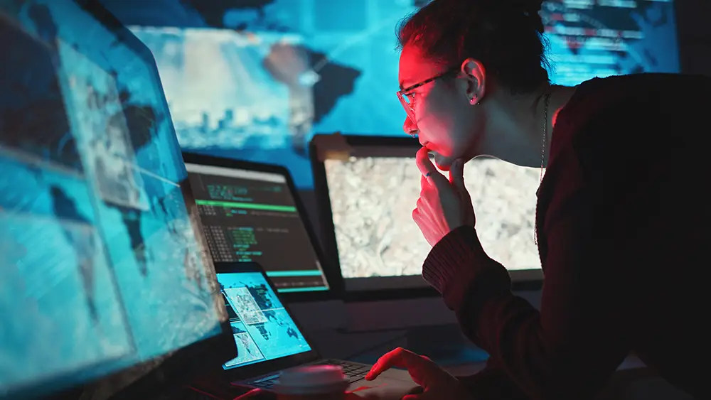 A person studies multiple computer monitors displaying data and maps in a dimly lit, high-tech global security operations center.