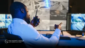 A man in a white shirt monitors multiple security screens and speaks into a walkie-talkie at a global security operations center (GSOC) control room.