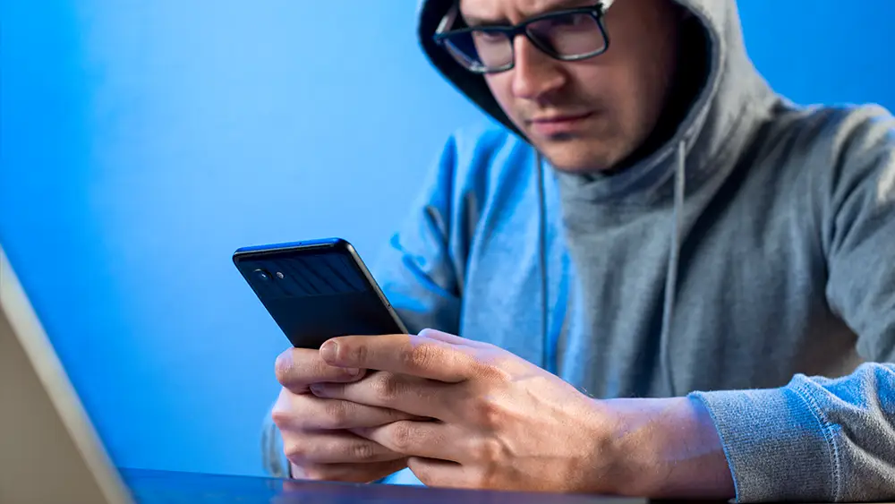 A person wearing glasses and a hoodie looks at a smartphone while sitting at a table with a laptop in front of a blue background.