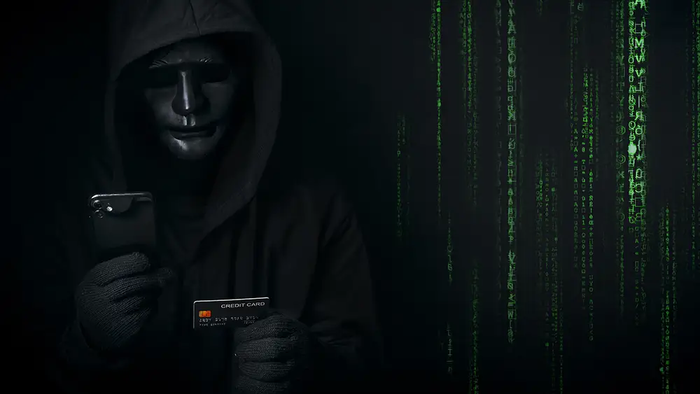 A person in a black hooded jacket and mask holds a smartphone and a credit card, with green digital code displayed on a dark background.