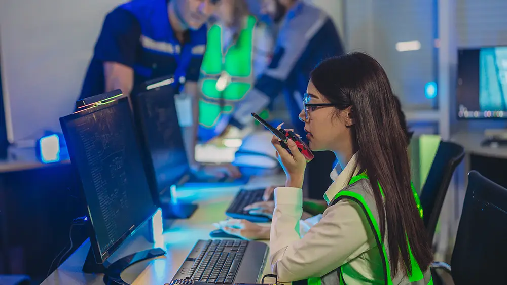 A woman wearing a reflective vest speaks into a walkie-talkie while sitting at a desk with computer monitors; two people stand in the background.