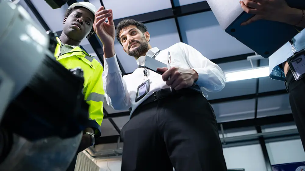 Two professionals, one in a safety jacket and hard hat, discuss equipment or machinery indoors, while another person holds a blue folder nearby.