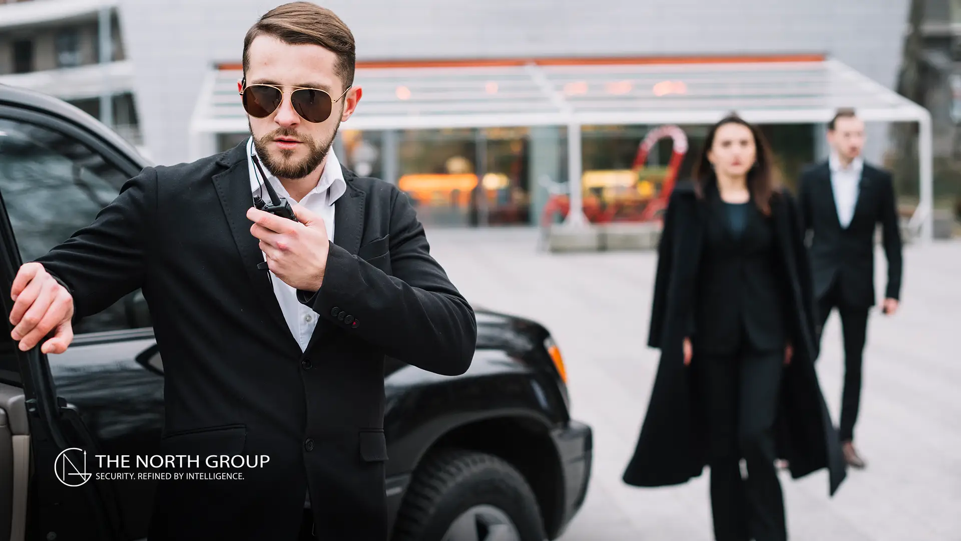 A security professional with sunglasses uses a walkie-talkie beside a black vehicle, while two people in business attire walk toward him in an urban setting.