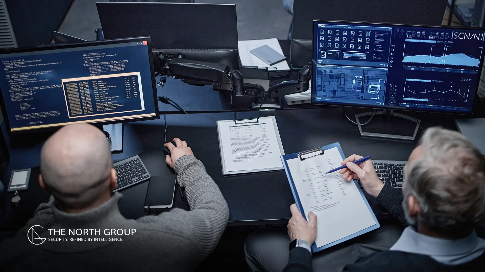 Two people sit at a desk with multiple computer monitors showing data and diagrams, one typing and the other writing on a clipboard with charts and notes.