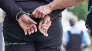 A person in a black shirt is handcuffed with hands behind their back while a law enforcement officer secures the cuffs. The North Group logo is visible in the bottom left corner.