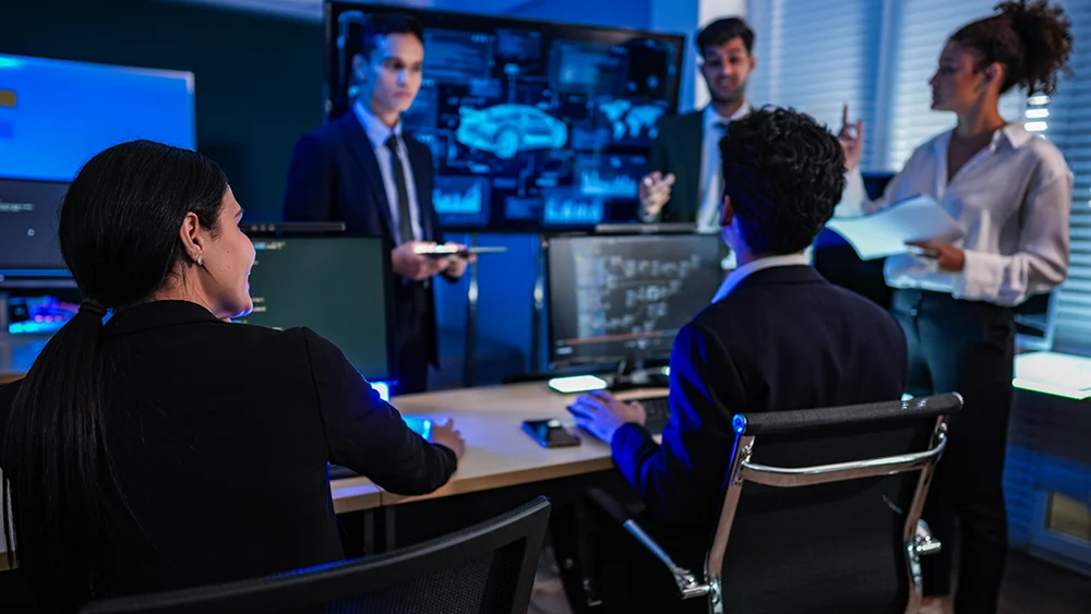 Five professionals in business attire have a discussion in a modern office with computers, charts, and digital screens displaying technical data and a car diagram.