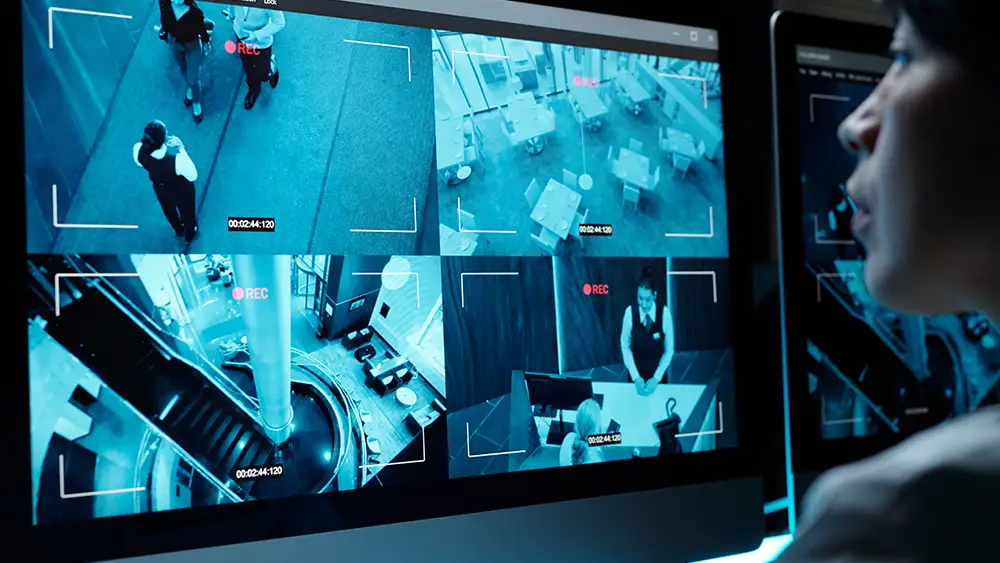 A person observes multiple live security camera feeds on a monitor, displaying various indoor locations and people in a building.