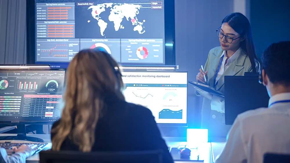 A woman stands and writes on a clipboard while two people sit at desks with multiple monitors displaying data charts and world maps in a control room.
