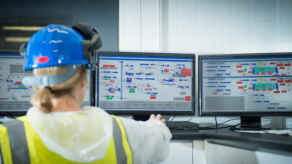 Person in safety gear works at a desk with three large monitors displaying complex data and control panels in an industrial control room.